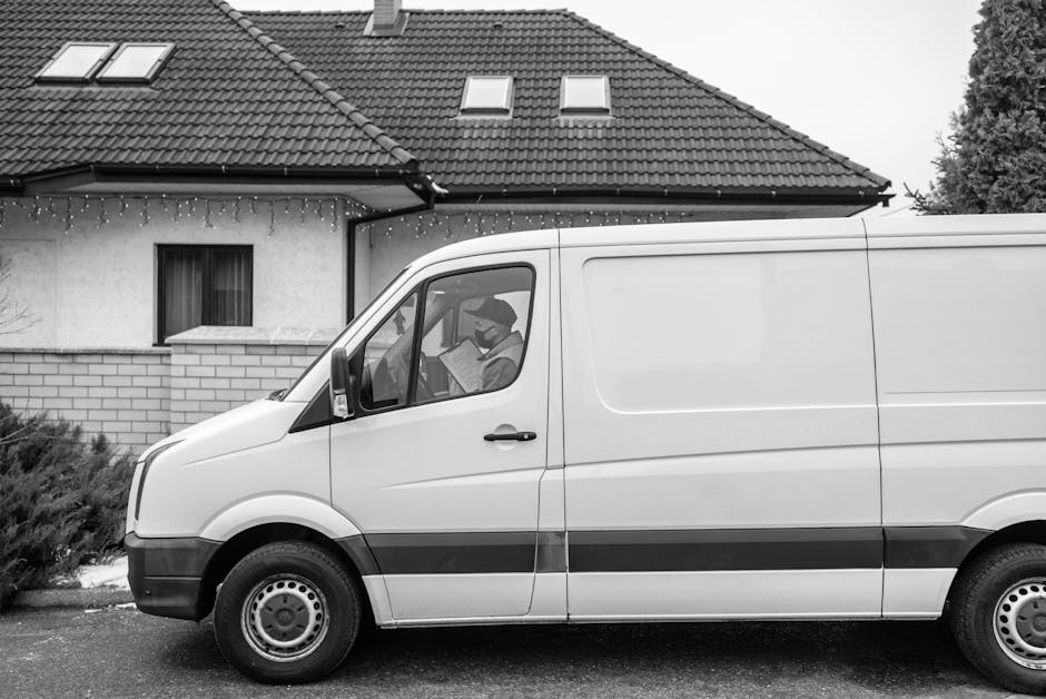 A man wearing a red and navy jacket and a blue beanie hat stands with arms crossed next to a grey delivery van with its side and rear doors open, revealing an empty interior with cardboard boxes and p